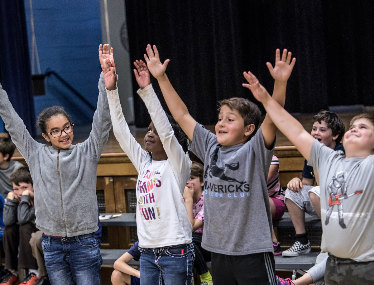 A group of young students with their arms raised at an in-school dance class.