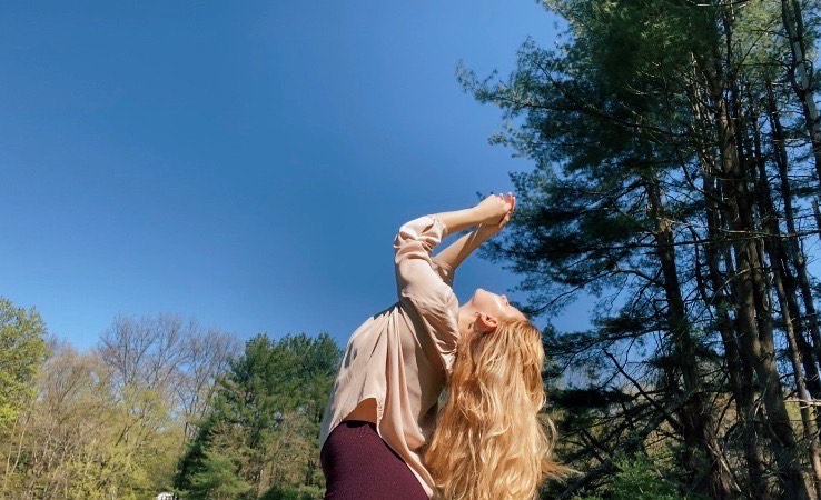 BFA Senior Madeleine Friedman, chest lifted to the blue sky, hands held together over head, green trees in the background