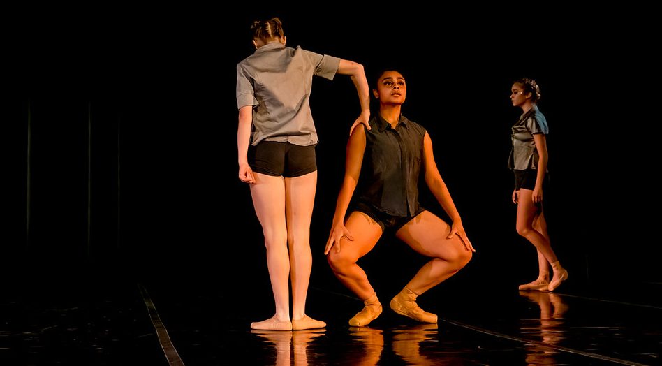 Amanda Harris dancing in her duet with fellow sophomore Brooke Sinton on the Angelico Hall Concert stage in the 2021 Fall Showcase. Another dancer enters onto the stage from behind. Amanda lowers into a grande plié as Brooke faces the back pressing down onto one of Amanda's shoulders.