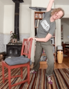 Teaching artist Erin Carper, dancing inside her home while using a chair as a barre, smiling to camera as she teaches a Dance for PD® at LINES class over Zoom