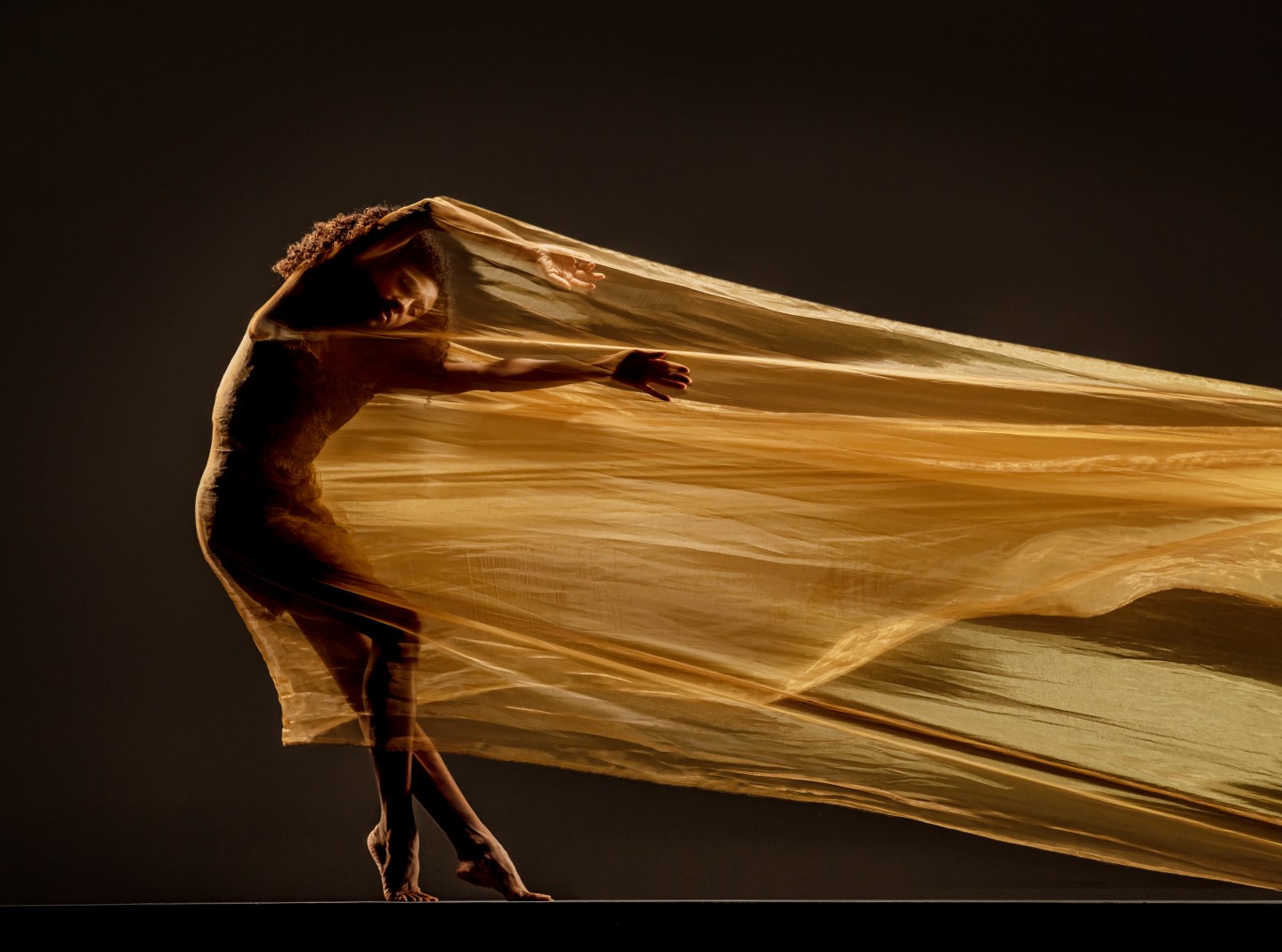 LINES Ballet company artist Adji Cissoko enclosed and intertwined into a flowing golden piece of fabric, stretching and pulling within the fabric