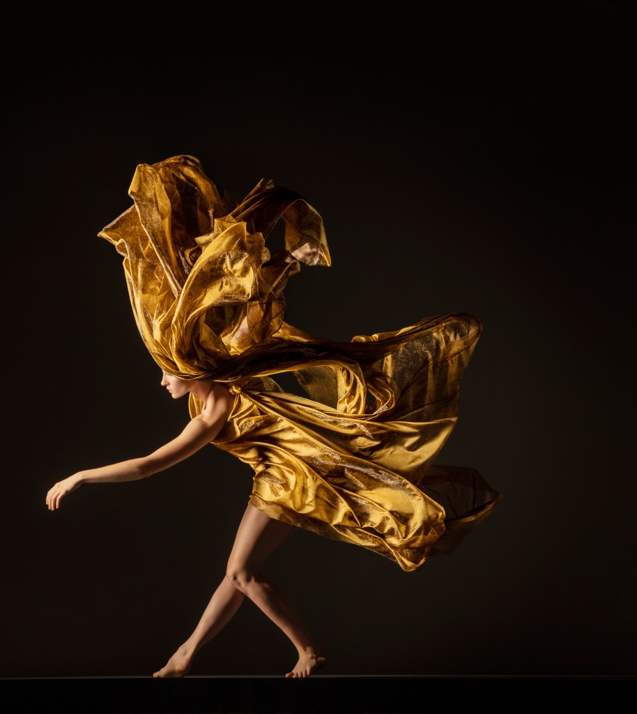 LINES Ballet Company Dancer Maya Harr extends one arm out while one knee bends and her golden, flowing dress wafts up in waves to cover half of her face