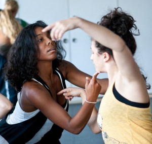 Sidra Bell dancing with a room of artists, Sidra is lightly holding a finger to one of the dancer’s chins while the dancer lightly holds a finger to Sidra’s heart