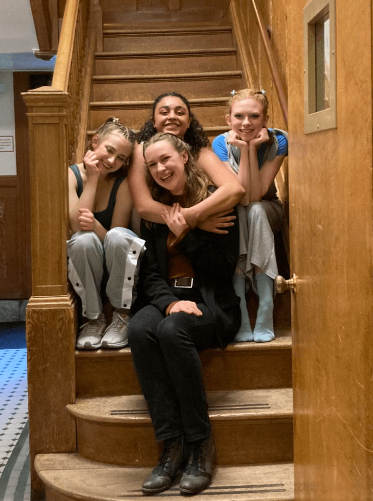 Amanda Harris, Brooke “Frank” Sinton, and Rowan Williams smiling and sitting with Hannah Woolfenden on a wooden staircase at Dominican University of California