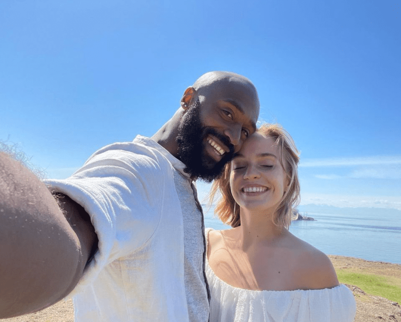 Babatunji taking a selfie with Charmaine Butcher with the ocean, sand, and blue sky in the background; Babatunji and Charmaine are smiling together