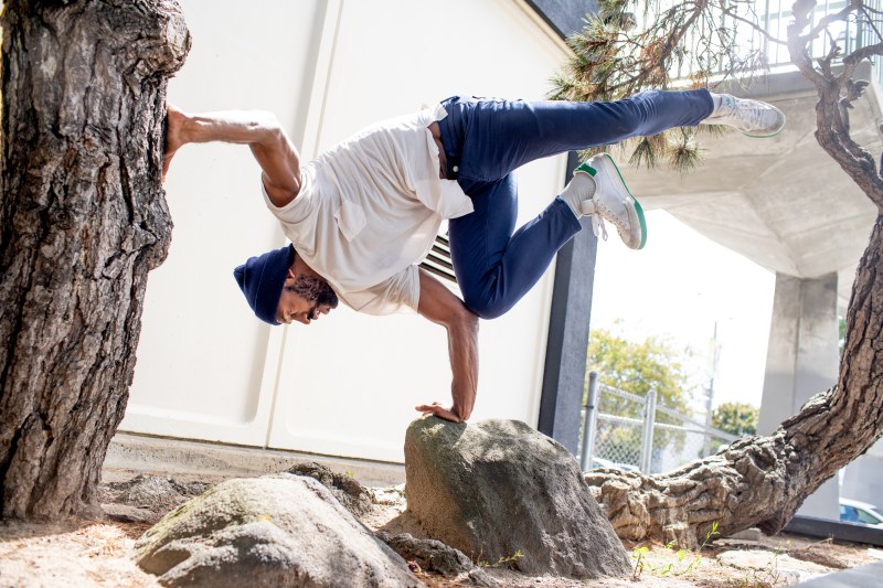 Babatunji in a freeze outdoors, one hand on a rock, the other pressing into a tree trunk; Babatunji is wearing a white T-shirt, blue pants, and white shoes with green accents