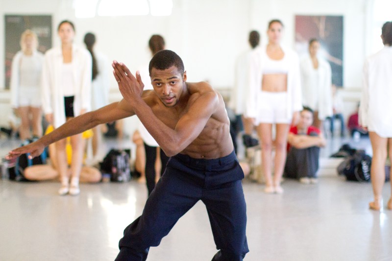 Babatunji as a LINES’ Training Program student dancing in a studio at LINES Dance Center; other Training Program students stand ready to perform in the background; observers sit on the floor
