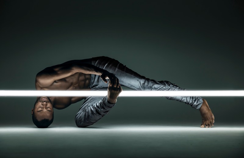 Alonzo King LINES Ballet dancer connects his leg and hand around a suspended glowing bar; he balances on the ground with his head, toes, and side of the knee resting on the floor with the rest of his body hovering above the ground; Babatunji’s eyes are closed