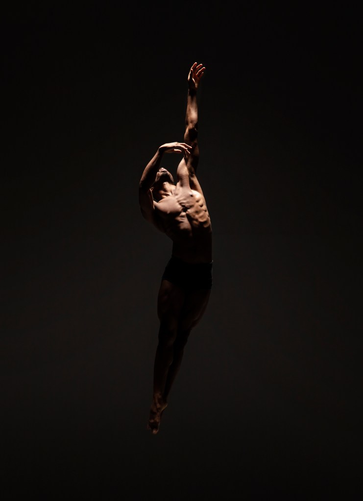 LINES Ballet company dancer Josh Francique jumping into the air against a black background; his head is tilted back while one arm extends up to the ceiling