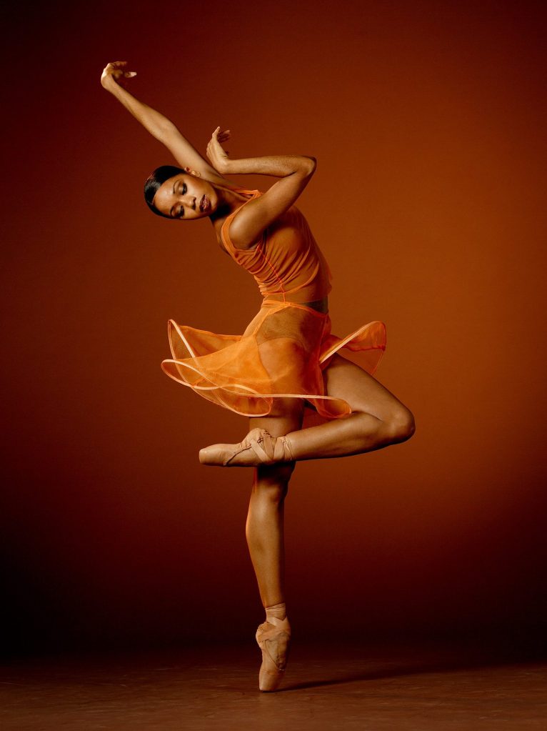 Former LINES Ballet dancer Aesha Ash in an orange skirt and leotard, dancing while balancing on one leg en pointe