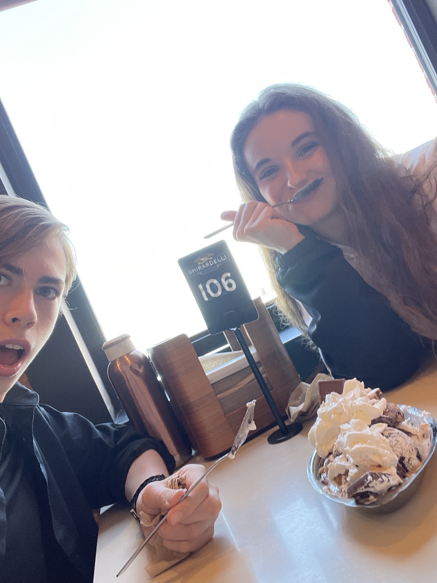 LINES Ballet Summer Program student Gabriel Warren with a fellow Summer Program student at Ghirardelli Square; both students are holding spoons and excited about an ice cream sundae on the table they're sitting at