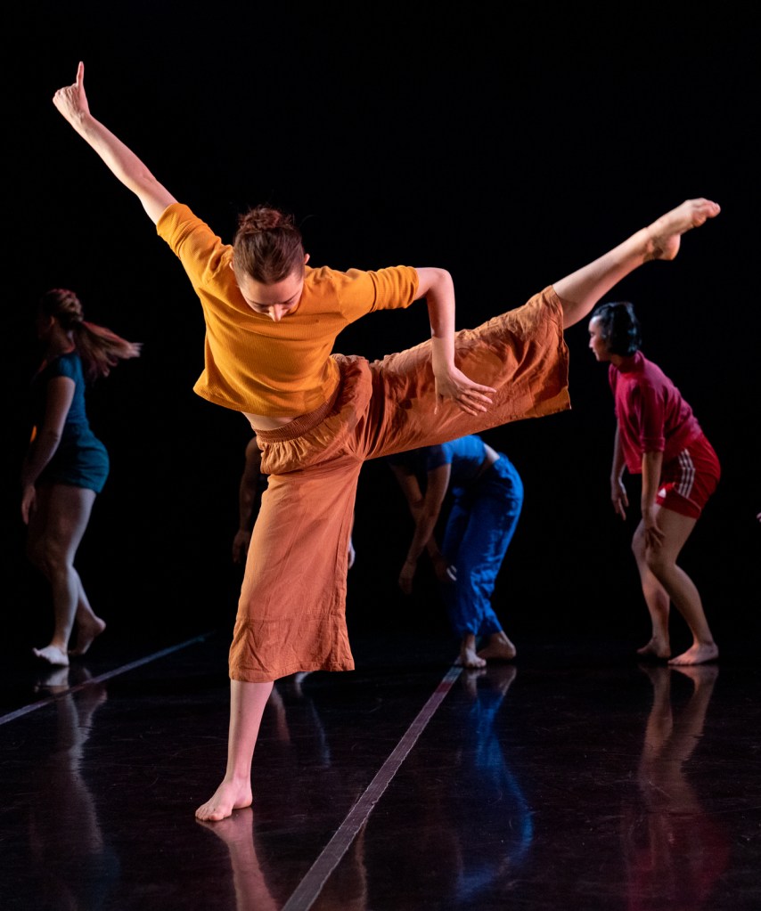 LINES Training Program students performing Katie Roy's piece "red fish blue fish" on stage; the focal dancer is wearing all orange, while half of the dancers in the back are wearing blue and the other half are wearing red