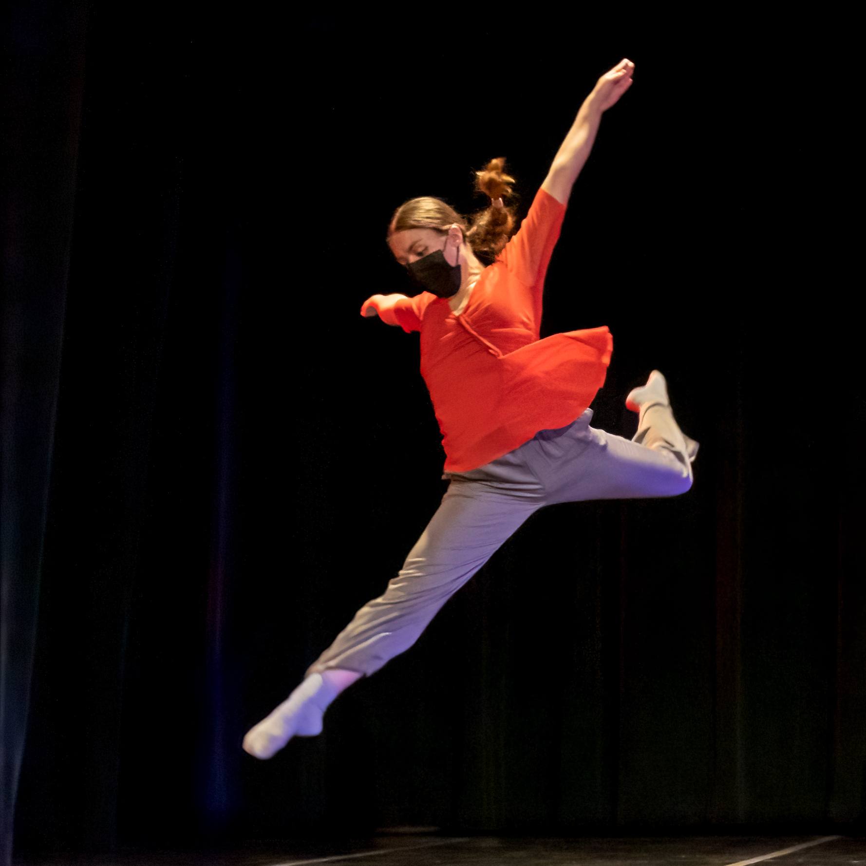 Emma Debski jumping while performing on stage at Angelico Concert Hall on Dominican University of California's campus; Emma is wearing a red shirt, gray pants, and a mask