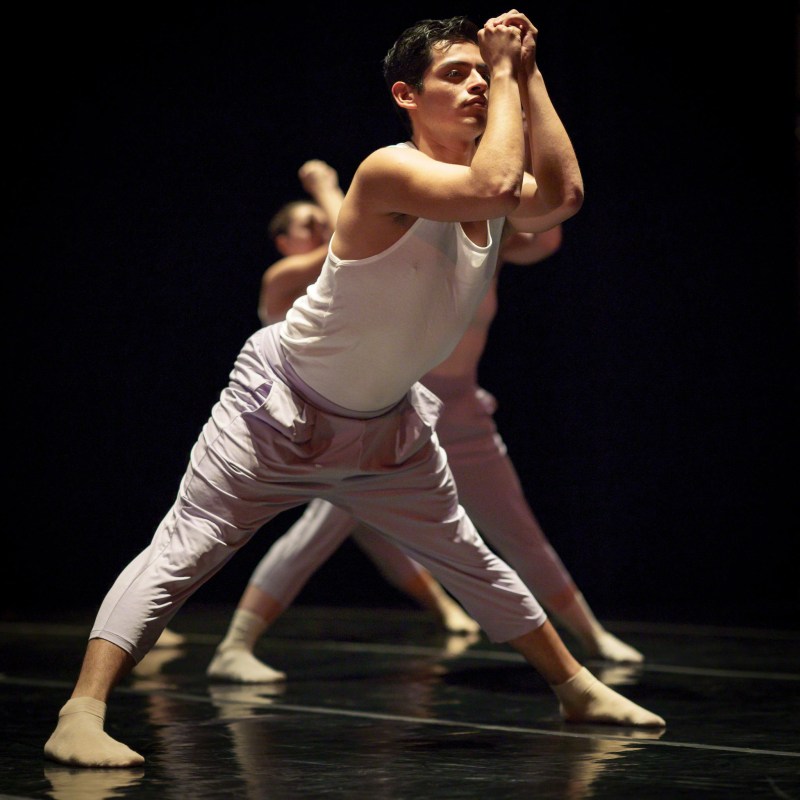 Héctor Jaime performing on stage at Angelico Concert Hall on Dominican University of California's campus; Héctor is standing in a wide second position with hands clasped together and held in front of the face; dancers are in the same position behind Héctor