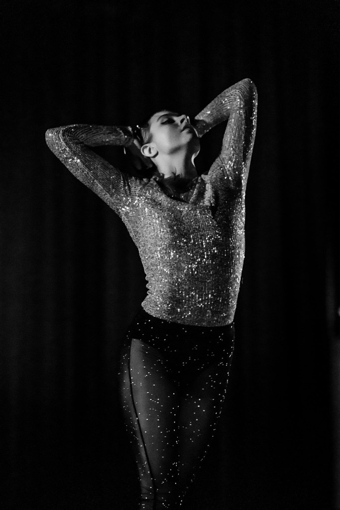 Emma Debski dancing on stage in a glittery top in sheer glittery black pants; Emma's back is bit arched with head held high with hands resting on the back of her head; the image is in black and white