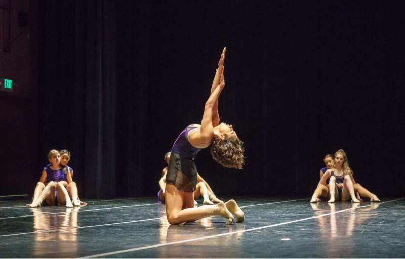 Zara Anwar performing on stage at Angelico Concert Hall on Dominican University of California's campus; Zara is resting on her knees, looking up with arms intertwined and extended above her head; her back is slightly arched; dancers Anouk Otsea and Gabriela Mendez and Ale Preciado and Marie Finely are sitting closely together in pairs on stage, watching Zara