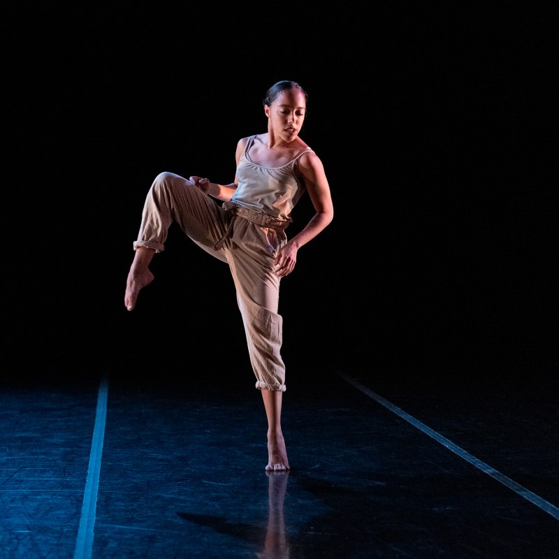 Giovana Sales dancing on stage in a Training Program Showcase; she is standing on relevé with the other leg in side attitude as she looks down over her opposite shoulder
