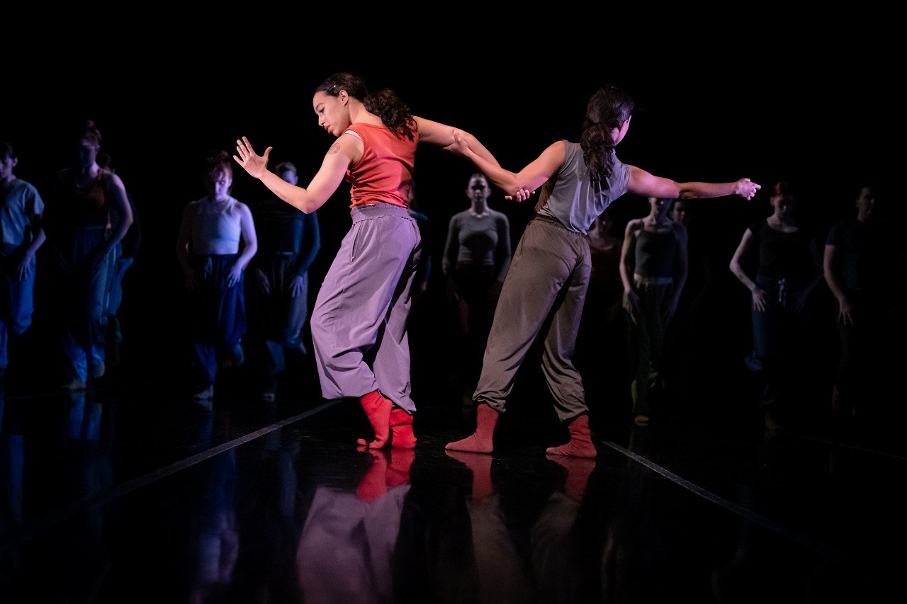 Giovana and Giulia Sales are performing on stage during a LINES' Training Program Showcase; both sisters are holding each other's elbow while facing different directions; other dancers in the background are facing front and walking forward