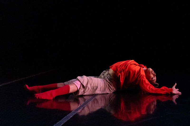 Giulia Sales performing a piece called "Her" by Anika Whetstone; Giulia is sitting on the ground with back arched towards the floor, eyes closed, and arm reaching back with fingers extended; her legs are extended out to the front with one knee slightly bent; she's wearing a red top, red socks, and rusty pink pants