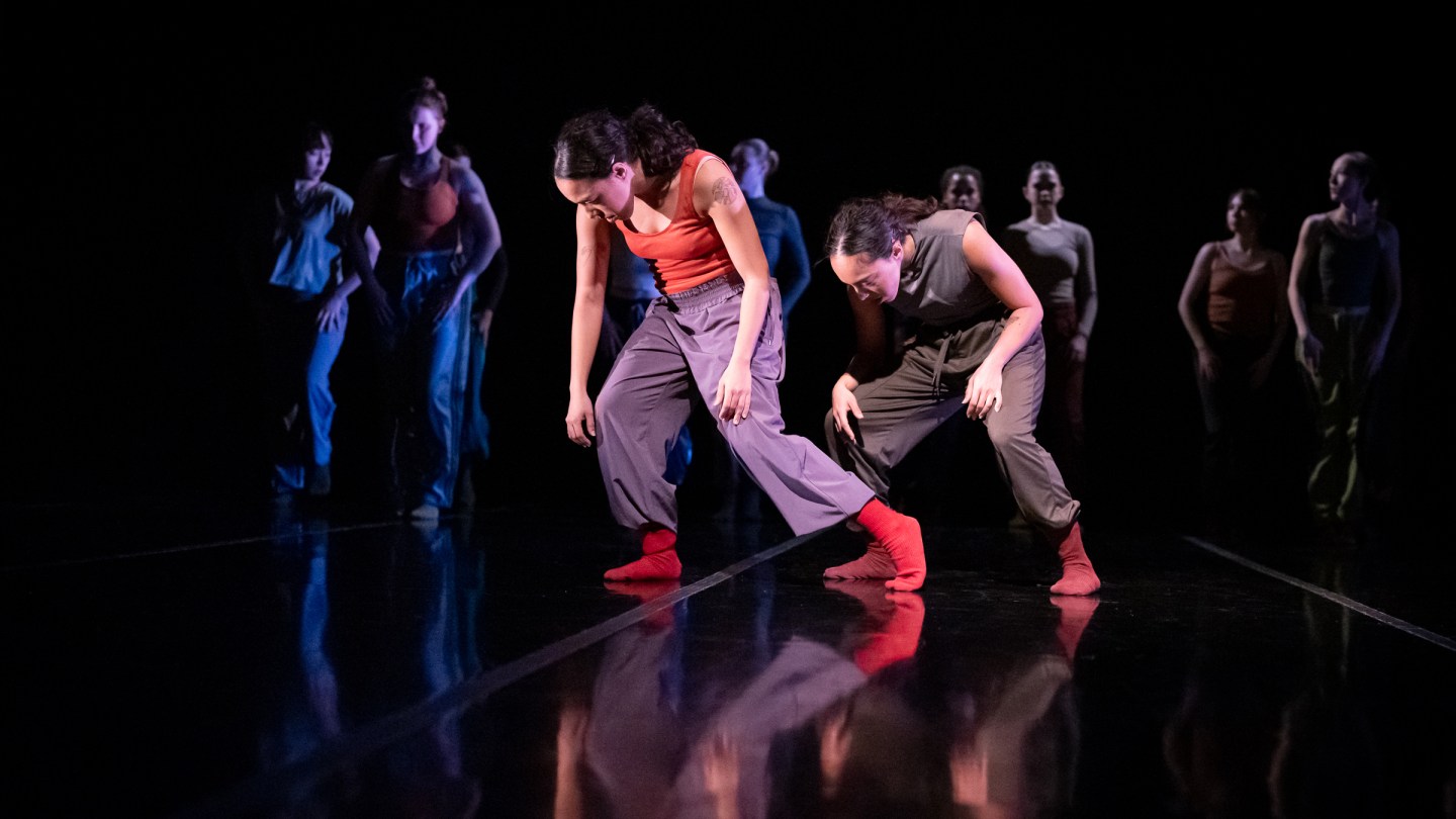 Giovana and Giulia Sales are performing on stage during a LINES' Training Program Showcase; both sisters are looking down, curling their backs forward with knees bent; other dancers in the background are facing front and walking forward