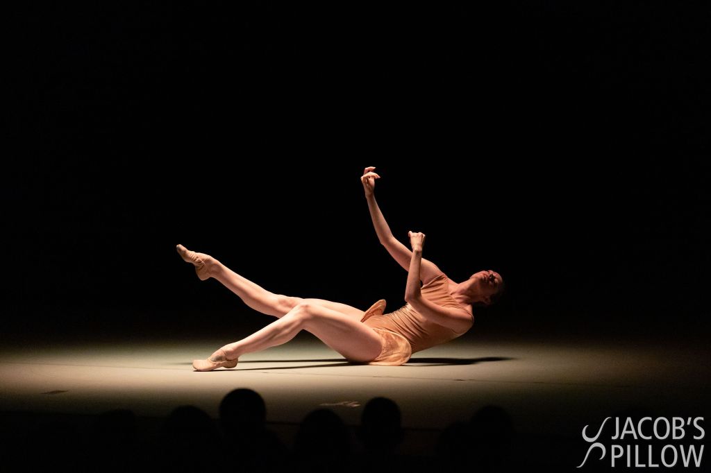 Alonzo King LINES Ballet Company Dancer Madeline DeVries performing on stage at Jacob’s Pillow; seated on the floor, Madeline suspends both of her legs up slightly off the ground, as her arms reach out and head leans back