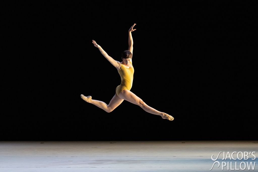 LINES Ballet Company Dancer Maya Harr wearing a yellow/golden leotard while jumping on stage, with one leg extended and the other bent at the knee; a Jacob’s Pillow logo is displayed in the bottom right corner