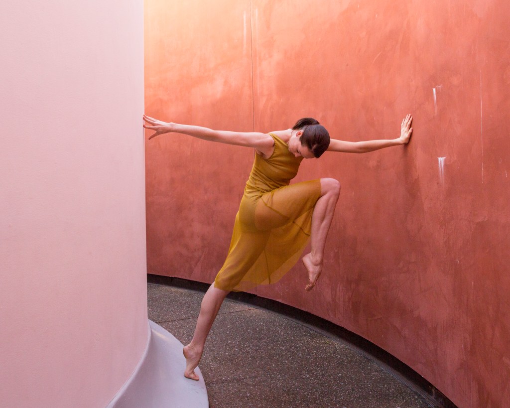 Alonzo King LINES Ballet Company Dancer Madeline DeVries standing on relevé on a wrap-around, raised step with the other leg in front attitude, leaning forward to rest her hand on the red/orange wall in front of her