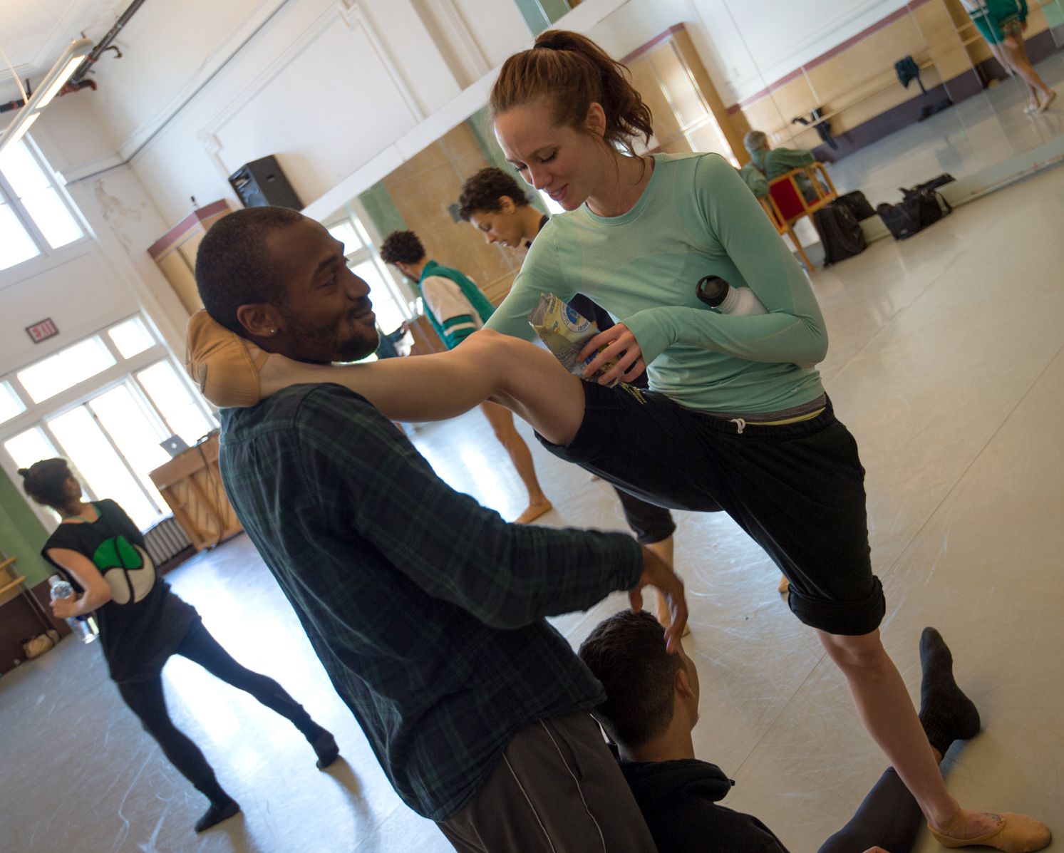 Alonzo King LINES Ballet Company Dancers Madeline DeVries and Babatunji together during a company rehearsal in studio 5; Madeline is resting her leg on Tunji’s shoulder, while Tunji’s stares at the snack she’s holding; both dancers are mid-laughter
