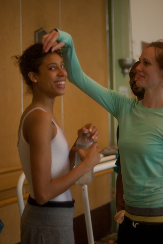 Alonzo King LINES Ballet Company Dancers Madeline DeVries and Adji Cissoko together during a company rehearsal in studio 5; Madeline is fixing Adji’s hair as Adji smiles at the camera