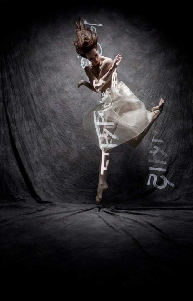 Alonzo King LINES Ballet Company Dancer Madeline DeVries jumping with arms crossed across her chest and hair flying up into the air; symbols made of light are projected onto her body and the back wall