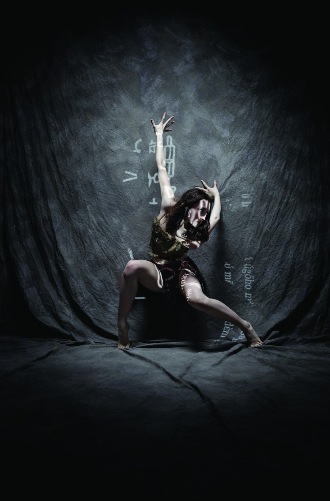 LINES Ballet Company Dancer Maya Harr crouching in a low position with arms extended behind her torso and fingertips touching the back wall; symbols created from light are projected onto Maya’s body and the back wall