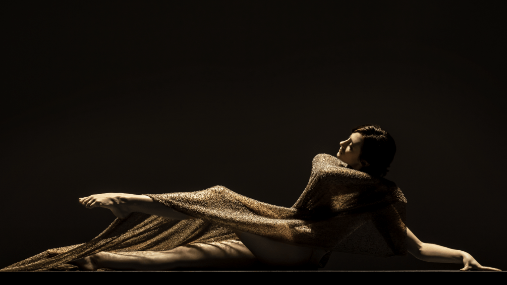LINES Ballet Company Dancer Maya Harr, sitting elegantly on the floor wrapped in a golden shimmery fabric, both legs are extended out with one on the floor, the other lifted slightly up and bent at the knee