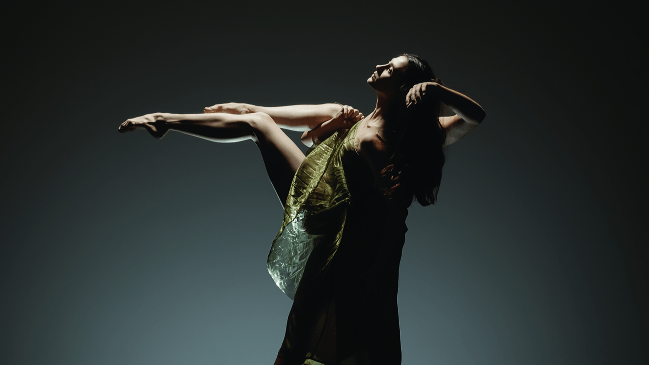 LINES Ballet Company Dancer Maya Harr is lifted into the air by a fellow company dancer who appears in silhouette behind her; Maya has her feet pointed and knees bent with one hand resting near the side of her face