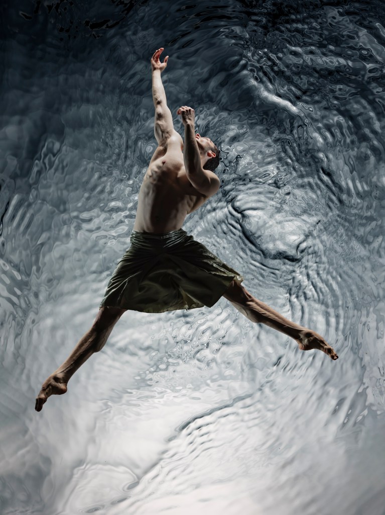 Company dancer Theo Duff-Grant jumps facing the sky in front of a rippling water backdrop