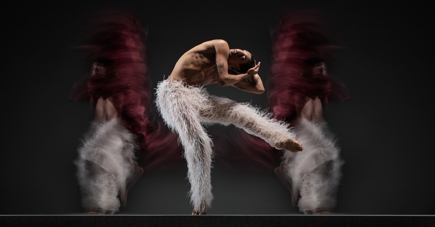 Company dancer Shuaib Elhassan bends forward with one leg up, wearing white feathered pants, in front of two blurred dancers on either side. Photo by RJ Muna