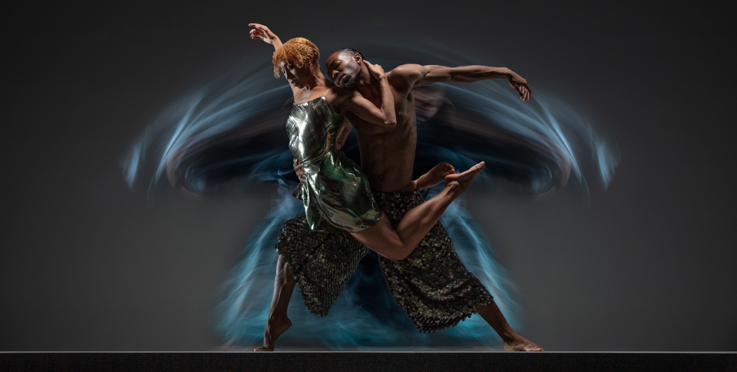 Company dancer Marusya Madubuko wraps her back around company dancer Joshua Francique, who supports her in a counterbalanced pose. Dancers in teal and green costumes.