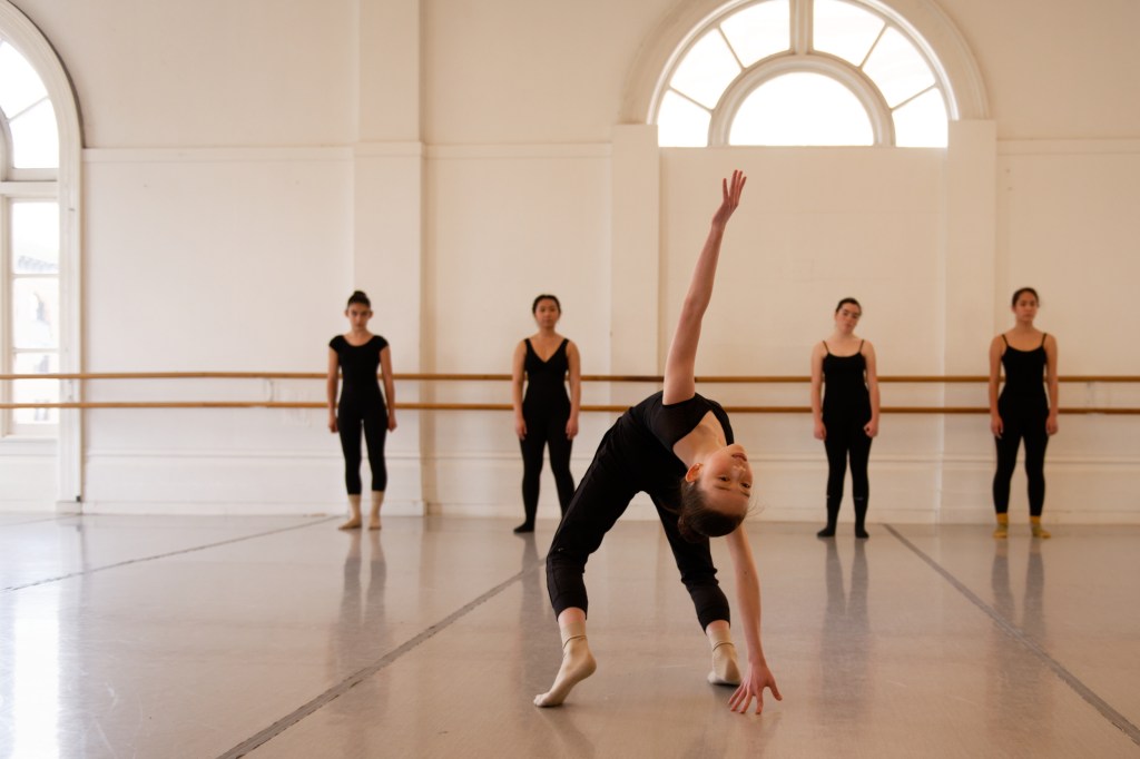 A young Education dancer bends backwards dramatically while classmates observe along the barre in the studio at LINES Dance Center.