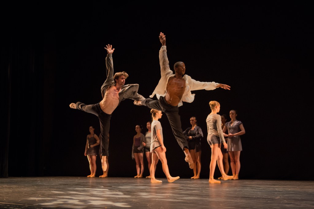 Two Summer Program dancers jump with technique and abandon, with a group of peers circling behind them.